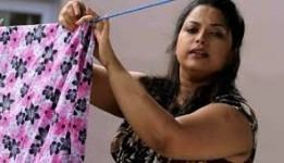 Suvarna Mathew hanging a floral blanket while looking down on something, an Indian actress with messy long hair half-tied, wearing a sleeveless blouse.