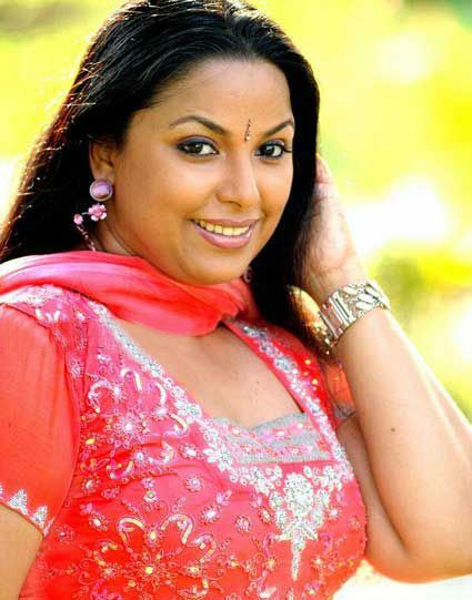 Suvarna Mathew smiling and posing with a bindi on her forehead and her left hand holding her hair with a silver bracelet on her wrist, purple dangling earrings, a red dupatta used as a scarf, and wearing a red beaded saree.