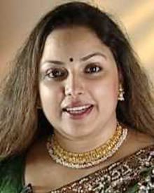 Suvarna Mathew smiling while talking to someone with a bindi on her forehead, an Indian actress with brown messy hair down, wearing gold earrings, gold and white layered necklaces, and a green brown-patterned saree.