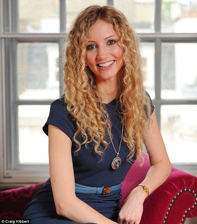 Suzannah Lipscomb smiling with curly blonde hair and a piercing on the left side of her nose, sitting on a red couch with a window behind. She has a lengthy pendant necklace, and a gold watch on her left wrist, wearing a blue belt with gold accent over a navy blue dress.