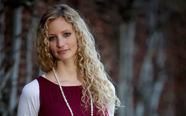 Suzannah Lipscomb smiling with curly blonde hair placed on her left shoulder and a nose piercing, wearing a lengthy white beaded necklace over a red sleeveless and a white sleeved blouse underneath.
