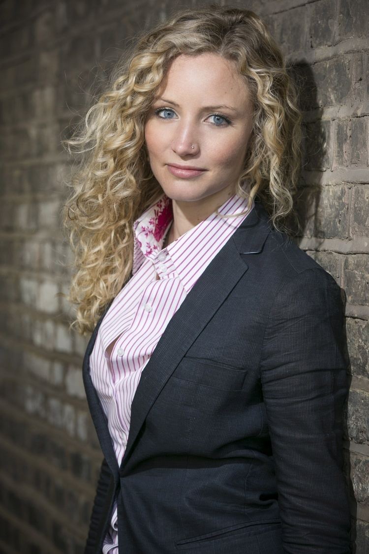 Suzannah Lipscomb smiling and leaning on a brick wall with curly blonde hair and a piercing on her left side of the nose, wearing a pink-striped collared shirt under a one-buttoned black coat.