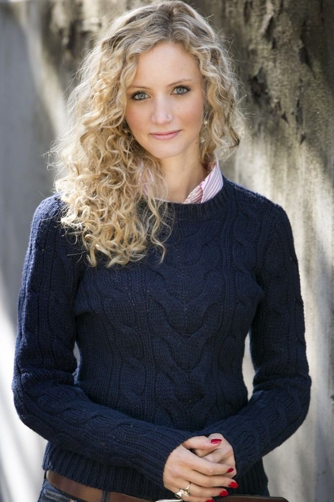 Suzannah Lipscomb smiling with a curly blonde hair placed on her right shoulder, and a piercing on her left side of the nose, holding her hands together with red polished nails, gold rings, and dangling earrings, wearing a pair of pants with a brown belt, and a pink striped shirt under a blue knitted jacket.
