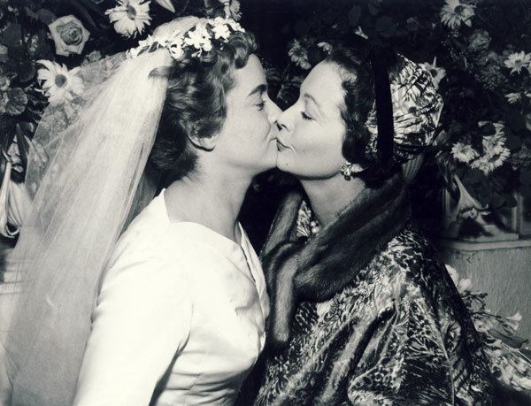 Suzanne Farrington and Vivien Leigh are kissing each other while closing their eyes at Suzanne’s wedding to Robin Farrington. Suzanne is wearing a white wedding gown and veil while Vivien is wearing earrings, a necklace, and a black fur dress.