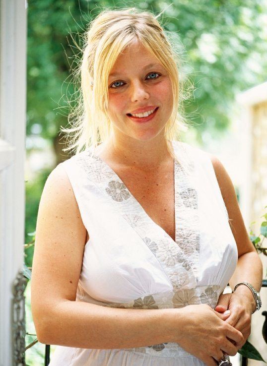 Suzanne Mizzi smiling, with blonde hair, and wearing a white sleeveless dress.