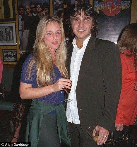 Suzanne Mizzi with long blonde hair, wearing a blue shirt while holding a glass of wine with Frank Camilleri smiling, with curly hair, and wearing a black suit and white long sleeves.