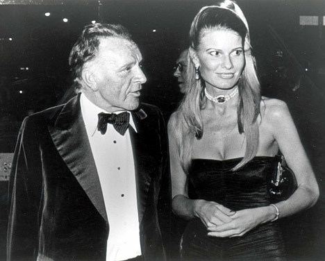 Richard Burton talking to someone while wearing a coat, long sleeve, and bow tie while Suzy Miller wearing a choker and tube dress