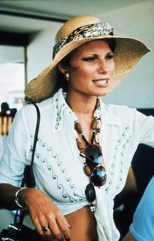 Suzy Miller talking to someone and carrying a black bag while wearing a brown hat, white long sleeve blouse, necklace, bracelet, and ring