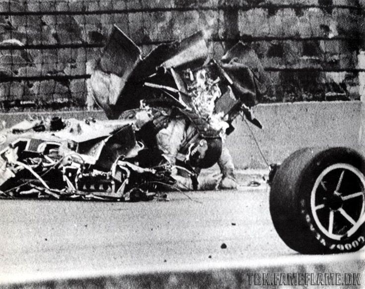 Swede Savage in a car crash, in the middle of a wrecked F1 car, facing down with his left hand on the ground, in a black and white, wearing a black helmet, at the right is a wheel of F1 race car.