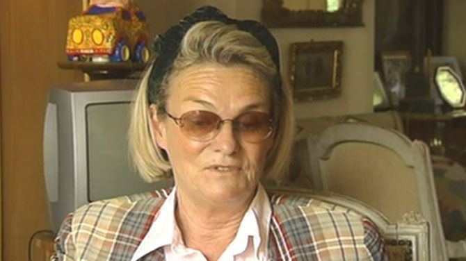 Sybille de Selys is talking while looking below with a TV and chair in the back, with short blonde hair, wearing a black headband, sunglasses, and a checkered blazer over a white polo shirt.