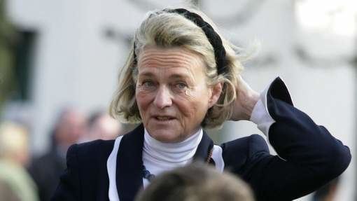 Sybille de Selys Longchamps is smiling while holding her short blonde hair, wearing a black headband and a black and white blazer over a white turtleneck top.