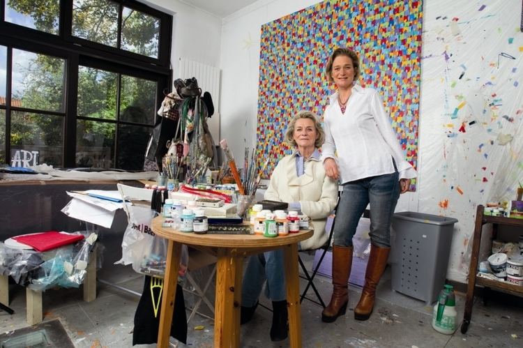 Sybille de Sélys Longchamps with a serious face while sitting in front of the table with art materials together with her daughter Delphine Boël smiling while standing next to her. Sybille with short hair, wearing a white coat over a light blue top, blue jeans, and black shoes while Delphine is wearing a necklace, a white long sleeve blouse, jeans, and brown boots.