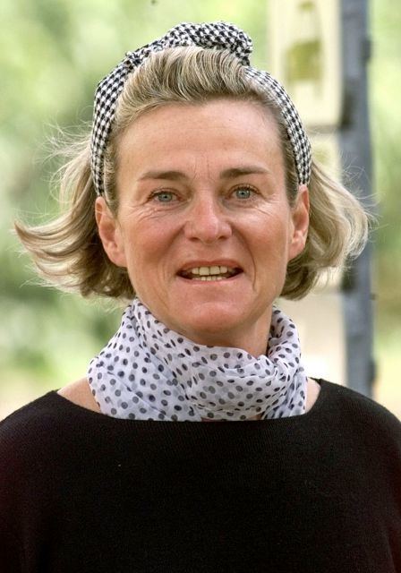 Sybille de Selys Longchamps smiling, with short blonde hair, wearing a black and white checkered headband, a polka dot scarf, and a black shirt.