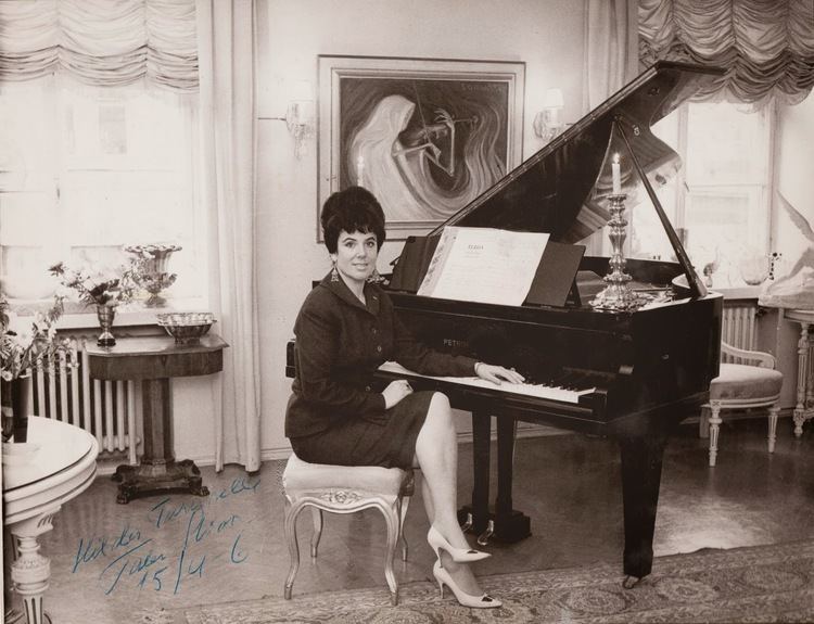 Tabe Slioor smiling while sitting beside a piano, wearing earrings, a modest dress, and white heels.