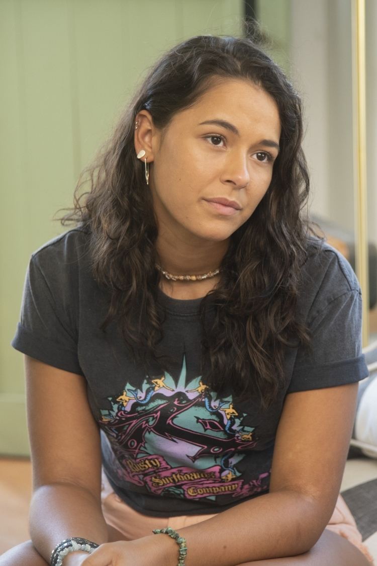 Tamala Shelton looking serious at something with her curly hair down and wearing a blouse, shorts, necklace, bracelets and ear piercings
