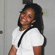 Tamika Huston smiling inside her house, sporting short hair and wearing a white shirt with a sling bag.