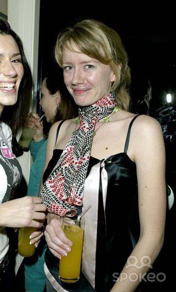 Tamzin Malleson and the woman beside her are smiling. Tamzin holding a glass of juice with blonde hair and bangs while wearing a black, white and red scarf, necklace, and a black and white spaghetti top