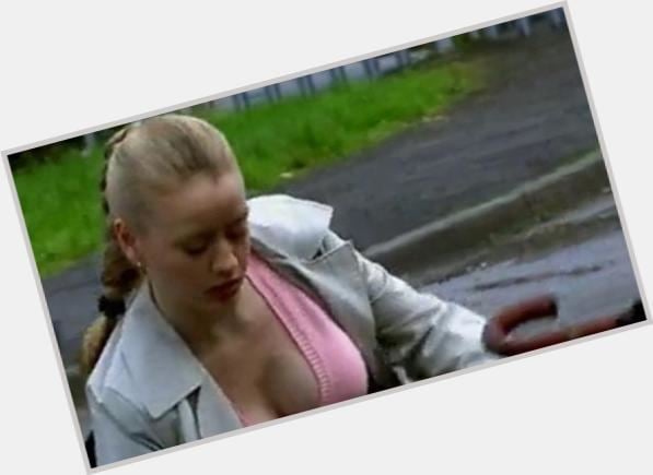 Tamzin Malleson looking down with braided hair while wearing a pink top under a gray coat that exposes her cleavage