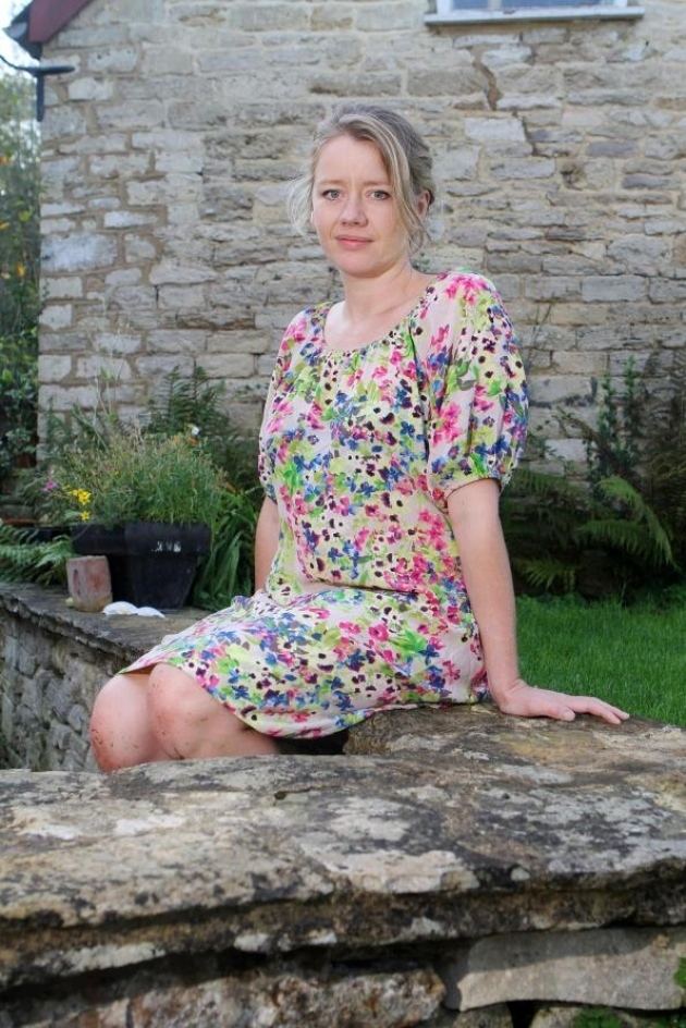 Tamzin Malleson with a tight-lipped smile while sitting on the wall with tied-up hair and plants behind her. Tamzin is wearing a colorful floral dress