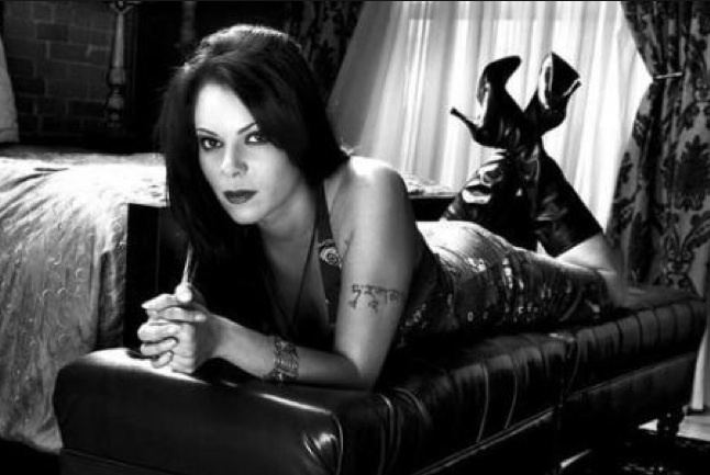 Tara Slone lying on a leather couch with a fierce look beside the bed in a brick wall and curtained window in the background. Her fingers were interlocked, with a tattoo, and a bracelet on her left arm, wearing a necklace and a printed halter dress with a pair of black leather heeled boots.