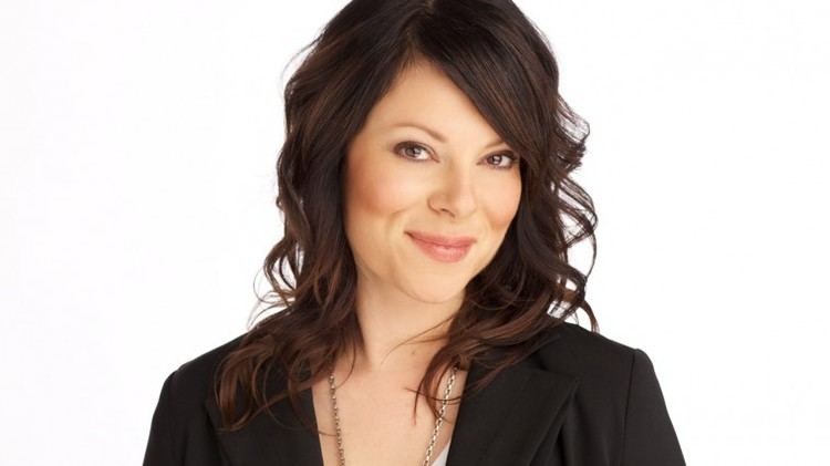 Tara Slone smiling in closed-lip on a white background. She has medium-length, brown and wavy hair, wearing a long, silver, chain necklace and a black coat over a white top.