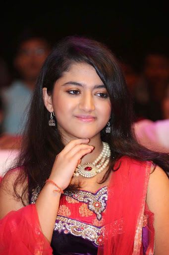 Shriya Sharma smiling while hand on her chin and wearing a red and blue dress, red dupatta, necklace, earrings, and bracelet