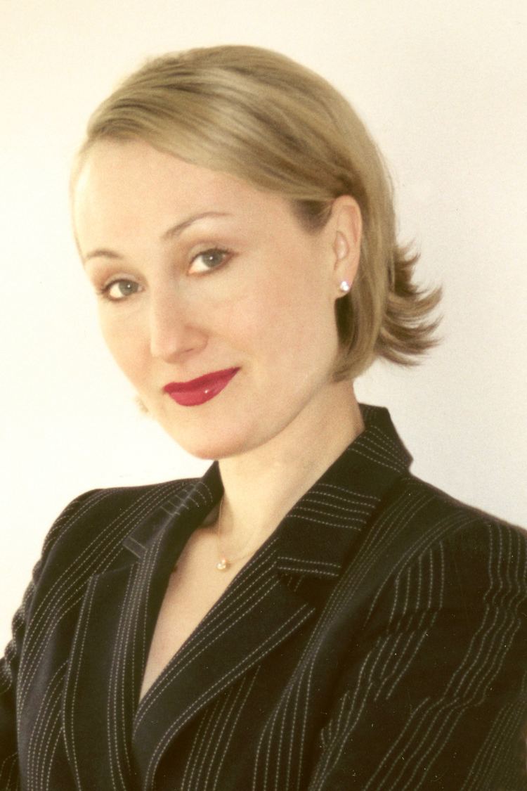 Tasha Kheiriddin with a tight-lipped smile, short blonde hair, wearing earrings, a necklace, and a black stripped blazer.