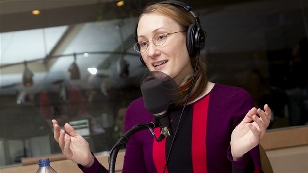 Tasha Kheiriddin is smiling while talking in front of a microphone, wearing a headphone, eyeglasses, and a multi-colored dress.