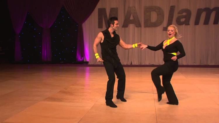 Tatiana Mollmann and Jordan Frisbee smiling, holding hands while dancing the West Coast Swing with white and violet curtains and violet led lights in the background. Tatiana in blonde hair and yellow bands on her neck chest and waist, wearing a pair of black swing shoes, a pair of black tights/leggings, and a skin-tight long sleeve top with a visible cleavage. Jordan has black hair, and yellow wristbands, and is wearing a pair of black swing shoes, black slacks, and a black sleeveless shirt.