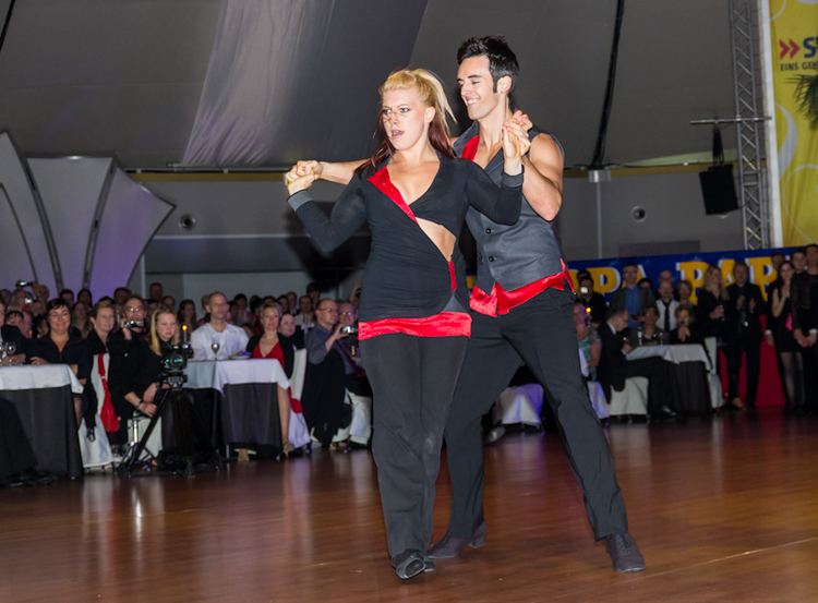 Tatiana Mollmann and Jordan Frisbee smiling, looking fierce, holding hands while dancing West Coast Swing with people, tables and chairs, a camera, and a speaker in the background. Tatiana has long blonde and brown hair tied up, wearing a pair of black swing shoes, black slacks, and a black and red long sleeve top with a visible cleavage and abs. Jordan has black hair, wearing a pair of black swing shoes, black slacks, and a black and red sleeveless top.