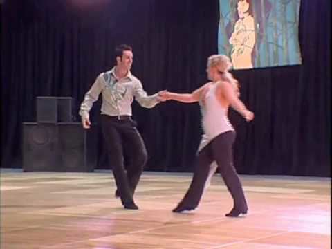 Jordan Frisbee smiling and holding the hand of Tatiana Mollmann while dancing West Coast Swing with black curtains, a banner, and speakers in the background. Jordan has black hair, wearing a pair of black swing shoes, a belt on his pair of black slacks, and a gray printed collared long sleeve. Tatiana has long blonde tied hair, wearing a pair of black swing shoes, black slacks, and a white backless overlapping top with a visible cleavage.