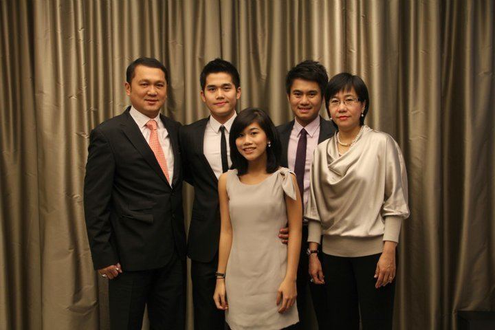 Tay Za smiling with his family while he is wearing a black coat, white long sleeves, black pants, and pink necktie