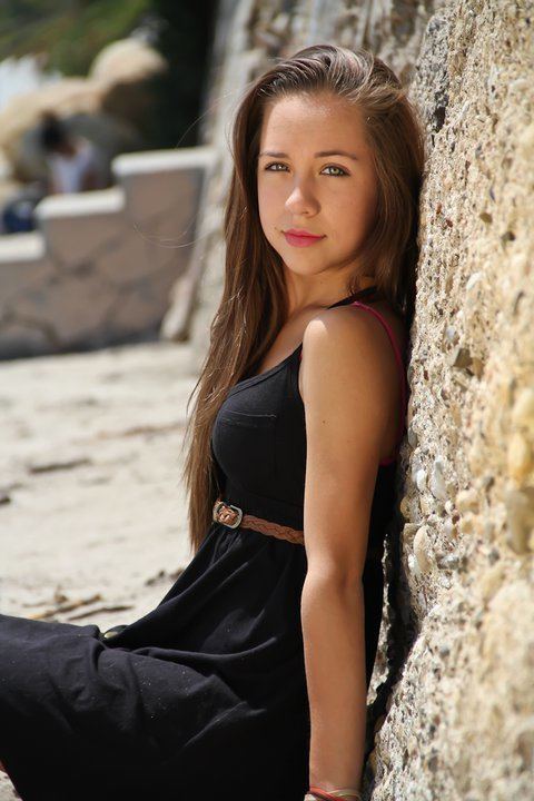 Taylor Atelian with a tight-lipped smile and leaning on the wall while wearing a bracelet and a black sleeveless dress with a brown belt and her pink bra strap is visible