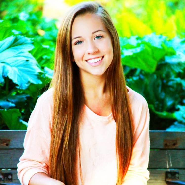 Taylor Atelian smiling and sitting on the bench with golden brown straight hair and plants behind her while wearing a light peach long sleeve shirt