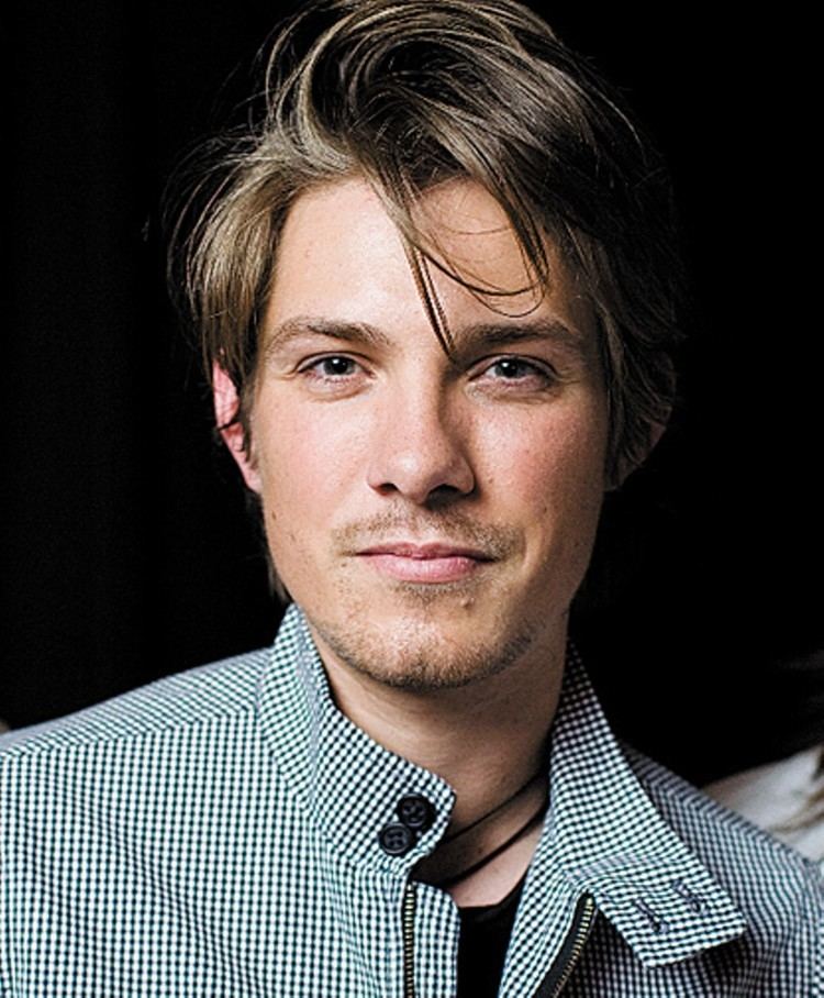 Taylor Hanson is smiling, has brown hair, gray eyes, visible moles under her left lips, cheek and neck, mustache and beard wearing a black necklace black shirt under a black and white checkered polo.