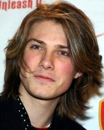 Taylor Hanson is smiling in front of white background poster with unleash written on top, has middle-length brown hair, gray eyes, visible moles under her left lips, cheek and neck, wearing a black top with silver beads.