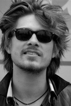 Taylor Hanson is smiling in black and white, mouth half open, has middle-length black hair, shaved mustache and  beard, wearing a black sunglasses, necklace with pendant and white polo under a black jacket.