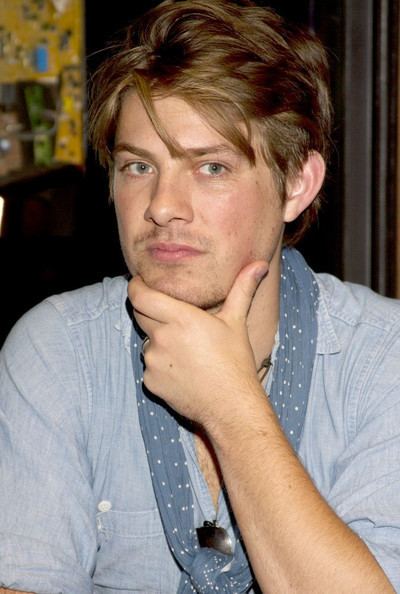 Taylor Hanson is serious, left hand under his chin, thumb under his left jaw while index and middle finger covering his chin, has brown hair, wearing a necklace with black pendant, dark blue white dotted scarf, and a blue denim long sleeve polo shirt.