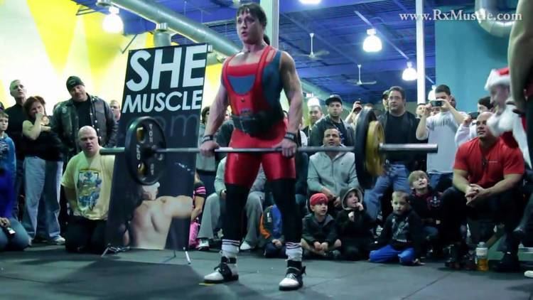 Tazzie Colomb holding a barbell and the people around is watching her while she is wearing a blue and red top and black and red pants