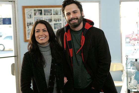 Michelle Borth and Luke Kirby are smiling in a scene from Tell Me You Love Me, a 2007 American cable television drama series. Michelle is wearing a black blazer over a gray turtleneck top while Luke has a beard and mustache, wearing a black and red hoodie jacket over a green shirt.