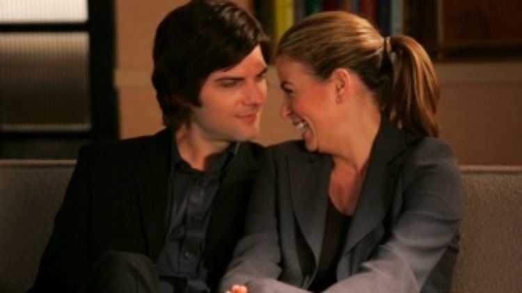 Adam Scott and Sonya Walger are smiling in a scene from Tell Me You Love Me, a 2007 American cable television drama series while sitting on a sofa. Adam is wearing a black coat over black long sleeves while Sonya is wearing a gray coat over a black shirt