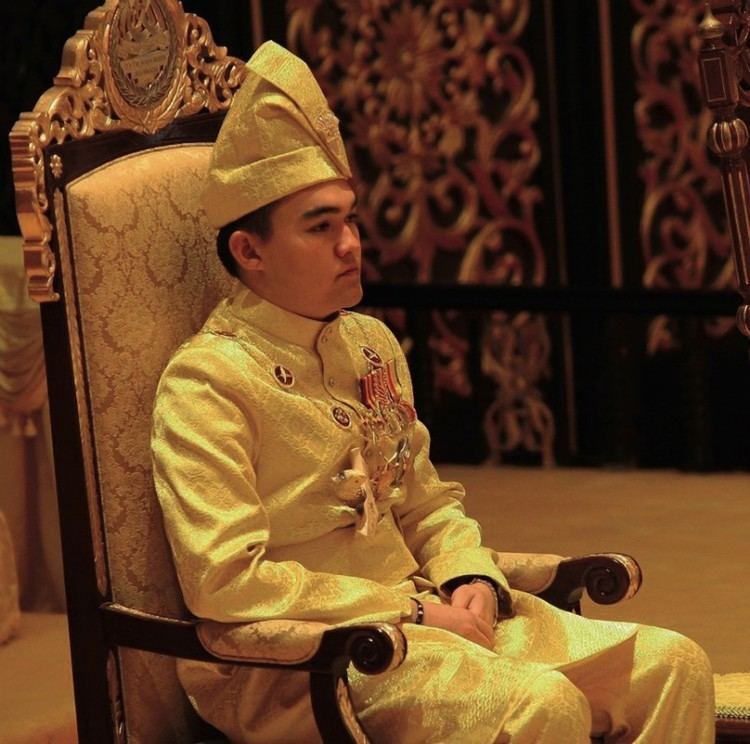 Tengku Amir Shah sitting on the throne while looking afar and wearing royal attire, yellow long sleeves with badges, pants, and hat