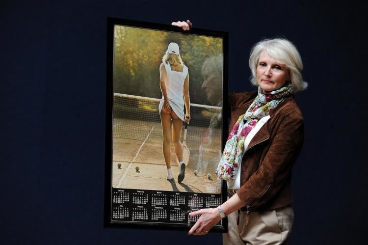 Fiona Walker holding the original copy of the Tennis Girl poster and she is wearing a brown coat, white inner blouse, scarf, and beige pants