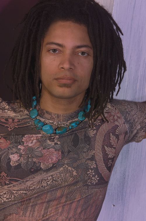 Terence Trent D'Arby with a poker face and twist hairstyle while wearing a pink and purple floral shirt and blue necklace