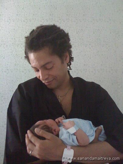 Terence Trent D'Arby smiling while looking at the newborn baby and he is wearing a black polo, necklace, and ring