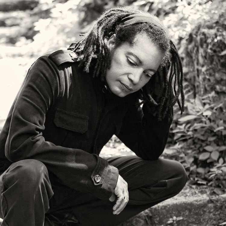 Terence Trent D'Arby looking at the ground with a twist hairstyle and wearing a headband, coat, and pants