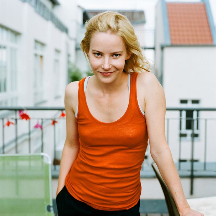 Teresa Weißbach with a tight-lipped smile, her right hand is in her pocket, with blonde wavy hair, and houses and railings in the background. Teresa wearing an orange sleeveless top with a white inner top and black pants
