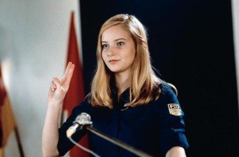 Teresa Weißbach as Miriam Sommer, in a scene from the 1999 German comedy film, Sonnenallee. Teresa with a tight-lipped smile, her two fingers are pointing upward, with blonde hair, there are flags in the background and a microphone in front of her. Teresa is wearing a dark blue blouse with a badge on her left arm