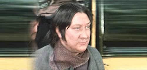 Terry Duggan with a tight-lipped smile while looking at something and wearing a gray coat and brown scarf