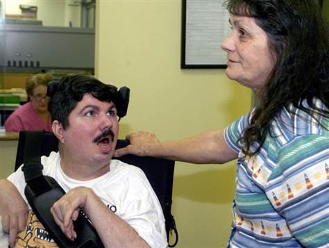 Terry Wallis sitting in a wheelchair and being accompanied by his mother for a doctors appointment with a black bag and wearing a white shirt.